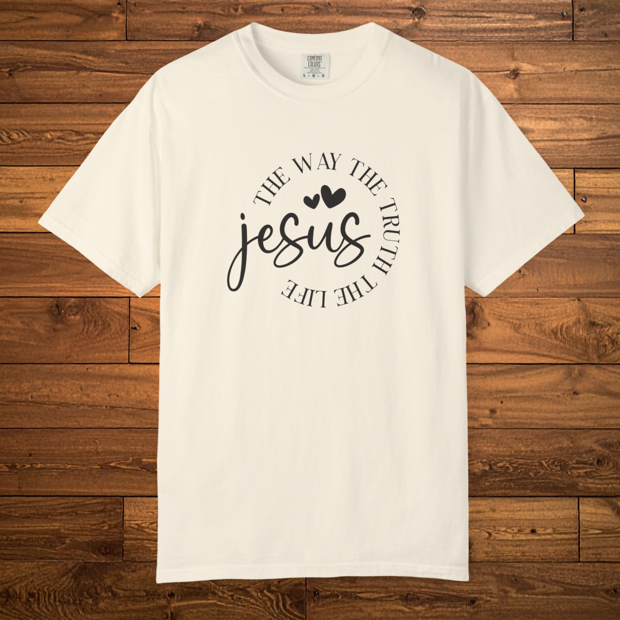 An off-white short-sleeve crewneck t-shirt with a black graphic print on the front reading 'THE WAY THE TRUTH THE LIFE' in an arc around the word 'jesus' in script, displayed flat against a brown wood plank background.