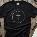 Black Christian graphic t-shirt laid flat, featuring a black cross surrounded by the message “Be who God has called you to be, not what the world wants to see” in a circular design.