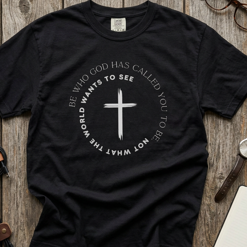 Black Christian graphic t-shirt laid flat, featuring a black cross surrounded by the message “Be who God has called you to be, not what the world wants to see” in a circular design.