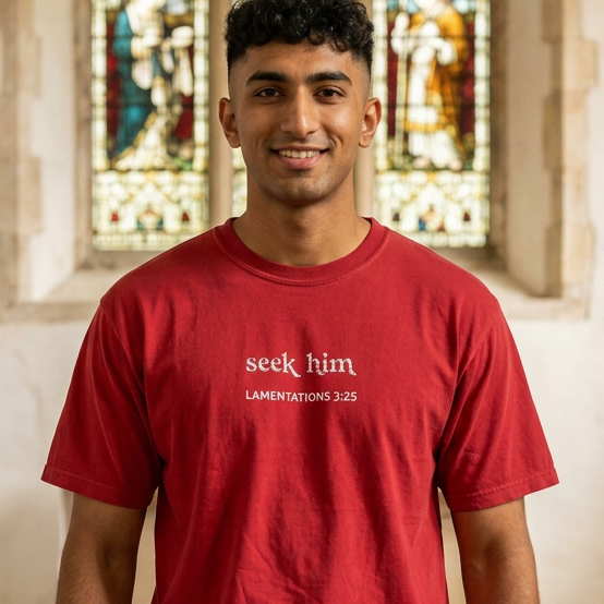 A man wearing a red crewneck t-shirt with the white embroidered text 'seek him' and 'LAMENTATIONS 3:25' stands in a stone corridor with stained glass windows in the background.