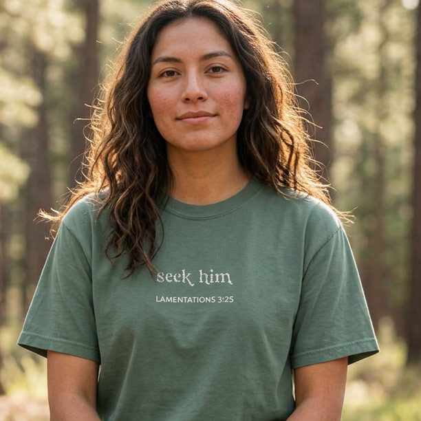 A woman sits cross-legged on a log in a forest, wearing a green t-shirt with the words 'seek him' and 'LAMENTATIONS 3:25' embroidered on the chest