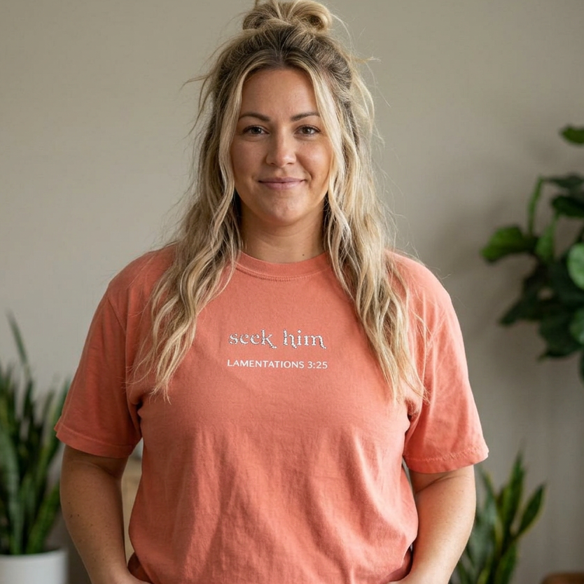 A woman with blonde hair in a messy bun stands indoors wearing a terracotta colored  t-shirt with the text 'seek him' and 'LAMENTATIONS 3:25' embroidered on the chest