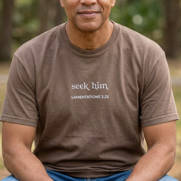 A man sits in a park, wearing a brown t-shirt with the text 'seek him LAMENTATIONS 3:25' embroidered in white