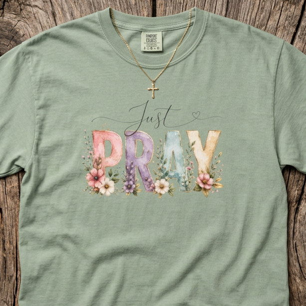 A sage green short-sleeve t-shirt with a graphic reading 'Just PRAY' in pastel floral letters, laid flat on a rustic wooden plank background.