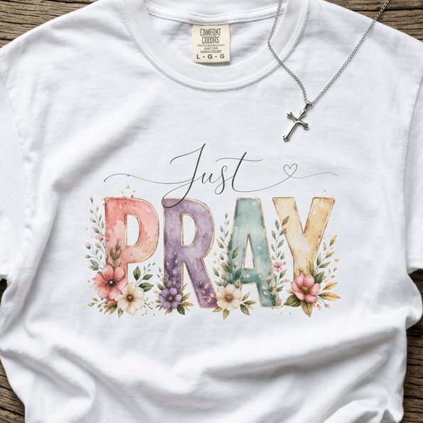 A white short-sleeve t-shirt with a colorful watercolor graphic reading 'Just PRAY' decorated with flowers, laid flat on a rustic wooden background. A silver cross necklace is draped over the collar.