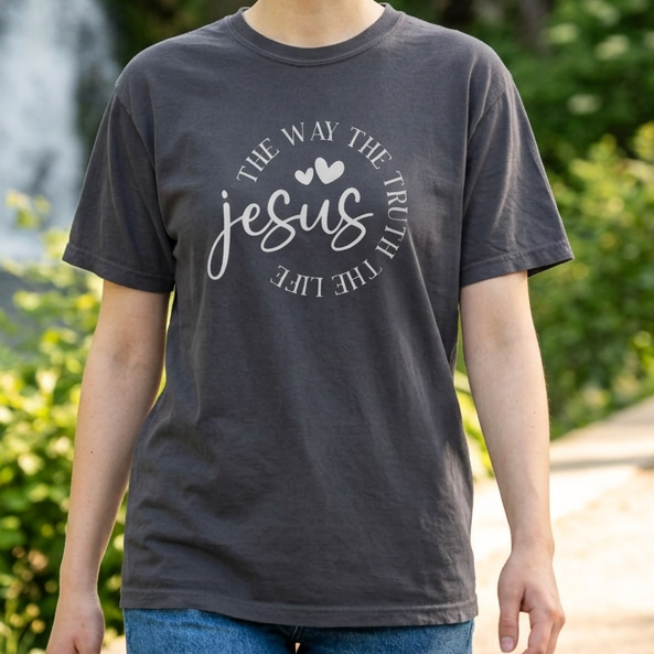 A smiling young woman with short blonde hair stands outdoors in front of a waterfall and green trees, wearing a dark charcoal gray graphic t-shirt with white text that reads 'The Way at the Cross Jesus is the Way of Life' and blue jeans.
