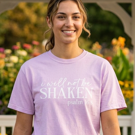 A woman wearing a light purple graphic t-shirt and blue jeans stands smiling in front of a white wooden gazebo with a garden background.