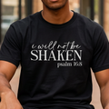 A man sitting on a bench in an urban setting wearing a black t-shirt with the text 'i will not be SHAKEN psalm 11:8' printed in white on the front.