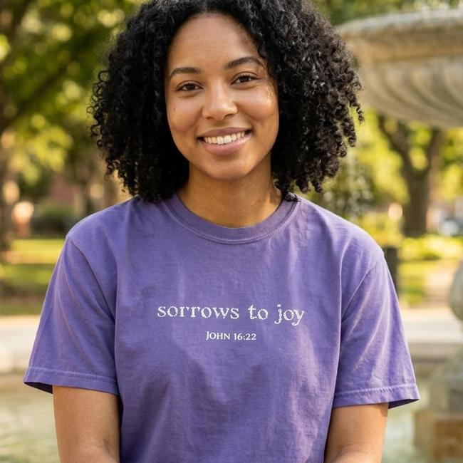 A smiling woman with curly black hair sits cross-legged outdoors in front of a fountain, wearing a purple short-sleeve T-shirt with the white printed text 'sorrows to joy' and 'JOHN 16:22', paired with blue jeans.