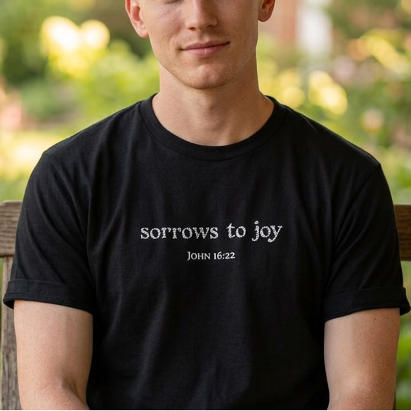 A man sitting on a wooden porch bench wearing a black t-shirt with the white printed text 'sorrows to joy' and 'John 16:22' below it, with a blurred garden background.