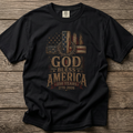 A black short-sleeve graphic t-shirt laid flat on a rustic wooden plank background. The shirt features a distressed patriotic graphic with a cross, American flag elements, and the text 'GOD BLESS AMERICA 250 YEARS 1776-2026'.