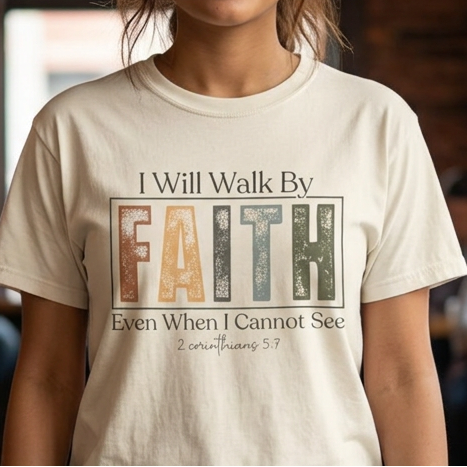 A woman stands in a cozy, blurred coffee shop wearing a cream-colored graphic t-shirt with the text 'I Will Walk By FAITH Even When I Cannot See' and 'Psalm 147:3'.