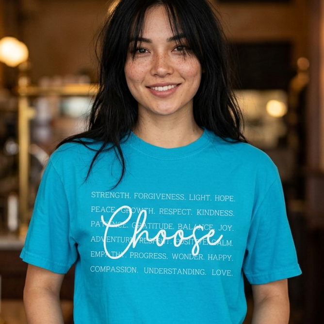 A woman with long dark hair stands in a warmly lit cafe or restaurant interior wearing a bright turquoise blue graphic T-shirt with white text that reads 'Choose' over a list of positive qualities. She is also wearing dark blue jeans, and the background shows blurred wooden furniture and warm lights.