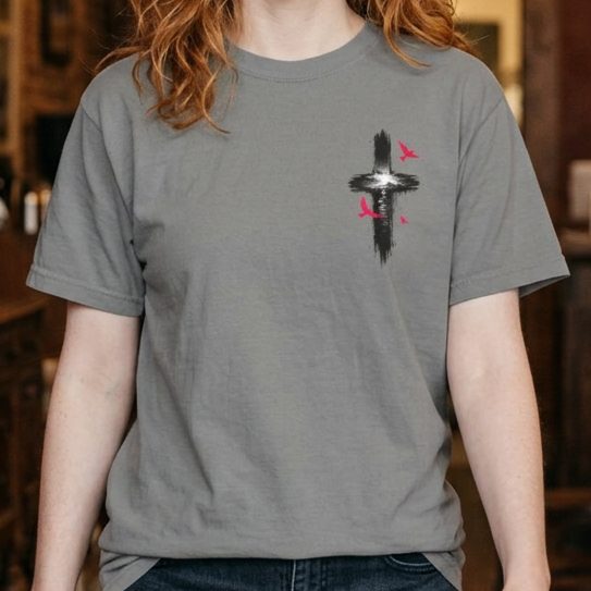 A smiling woman with red hair stands in a rustic coffee shop wearing a heather gray t-shirt with a small graphic on the left chest, paired with dark blue jeans.