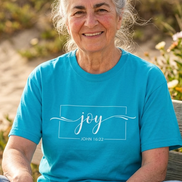 An older woman with gray hair sits cross-legged on a wooden beach bench wearing a bright turquoise blue t-shirt with 'joy JOHN 15:13' printed in white on the chest, set against a blurred sandy dune background.