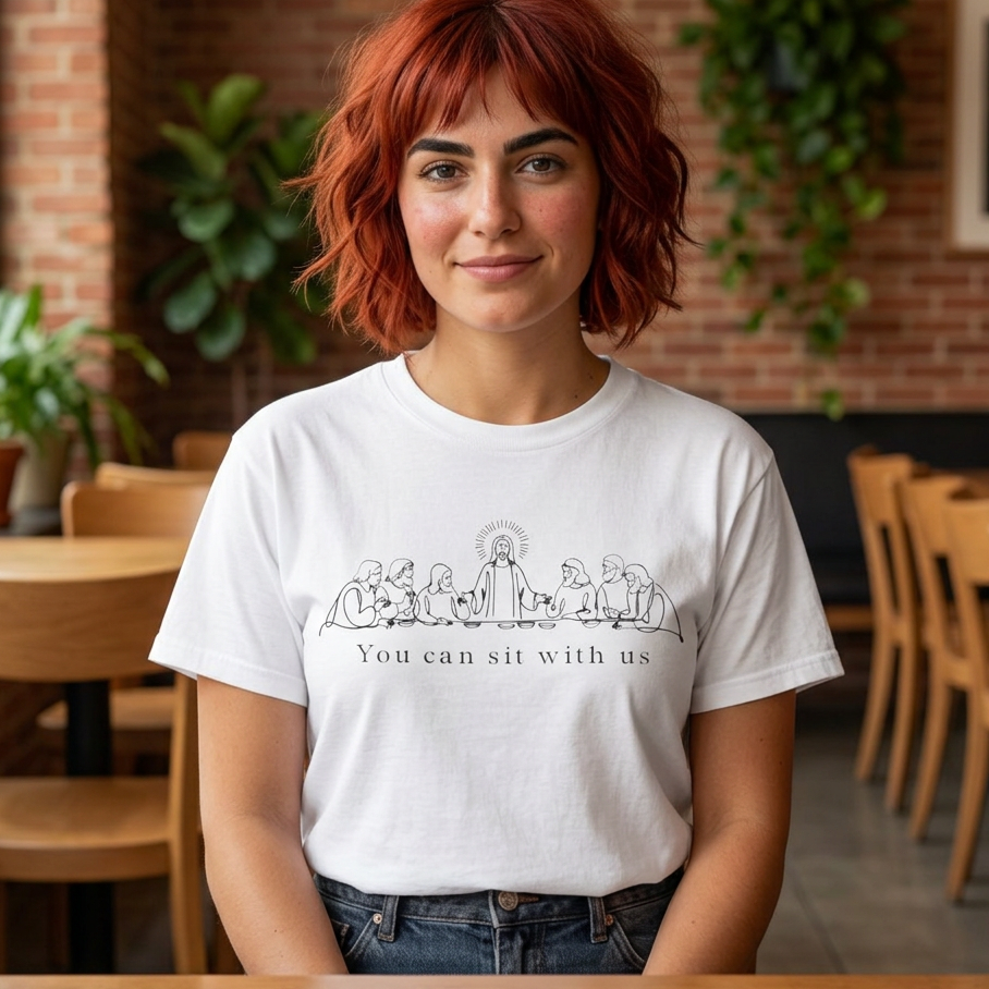 A woman with short red hair wearing a white graphic t-shirt with a black line art illustration of Jesus and the text 'You can sit with us', standing in a cafe with a brick wall background.