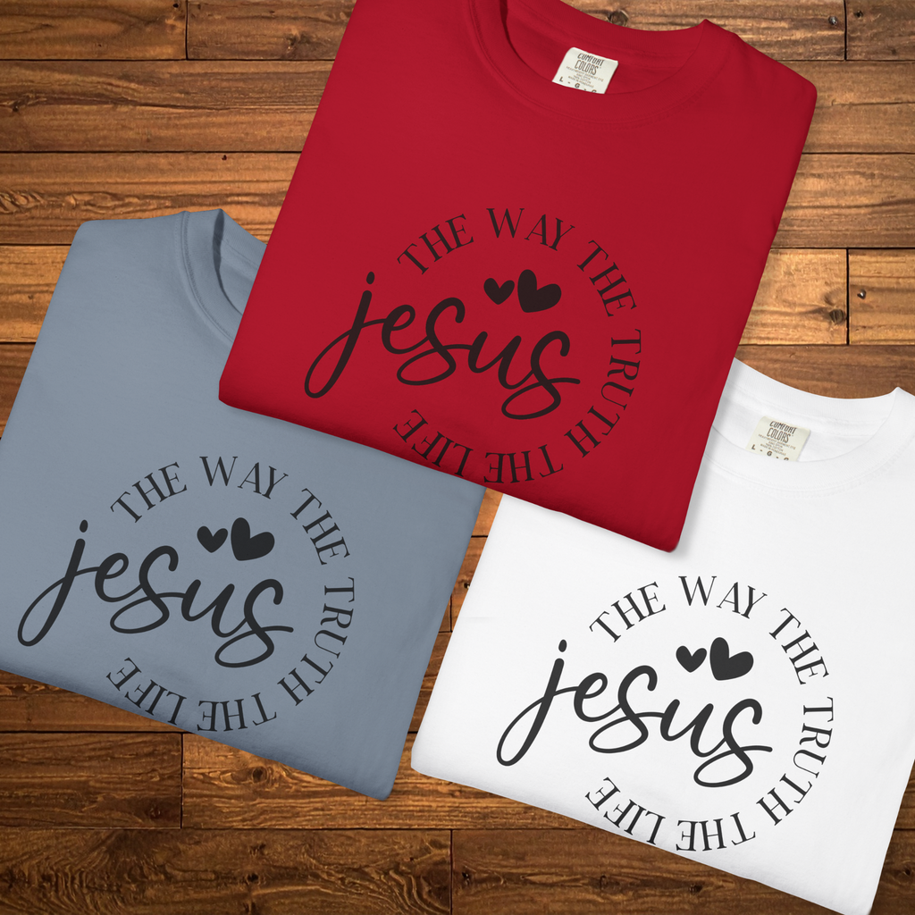 Three folded T-shirts in red, light blue, and white are arranged on a wooden surface. Each shirt has a black printed graphic with the words 'THE WAY THE TRUTH THE LIFE' in a circular arrangement around the word 'jesus' in script, with two small hearts above it.