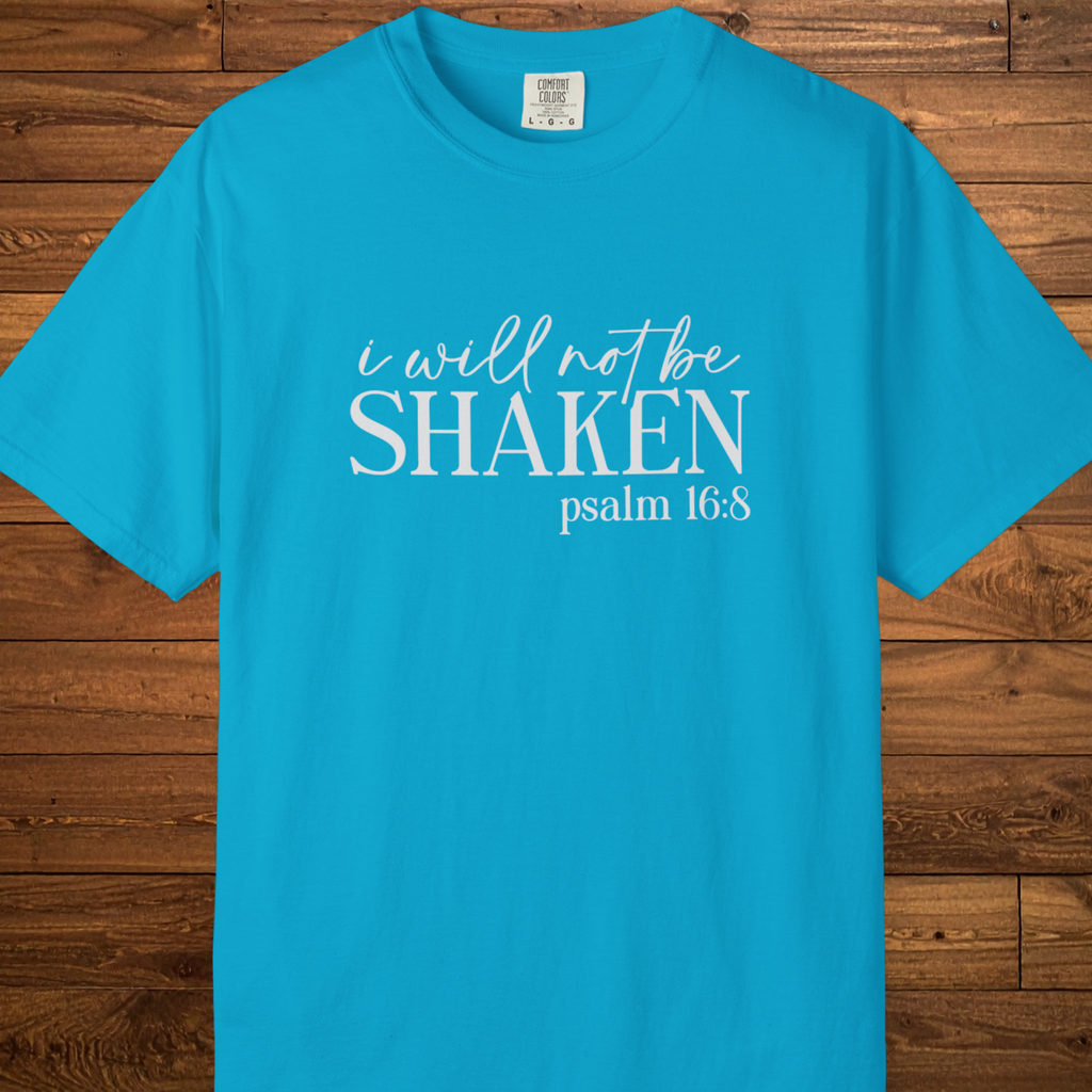 A bright turquoise blue short-sleeve t-shirt with white printed text reading 'i will not be SHAKEN psalm 16:8' laid flat against a brown wood plank background.
