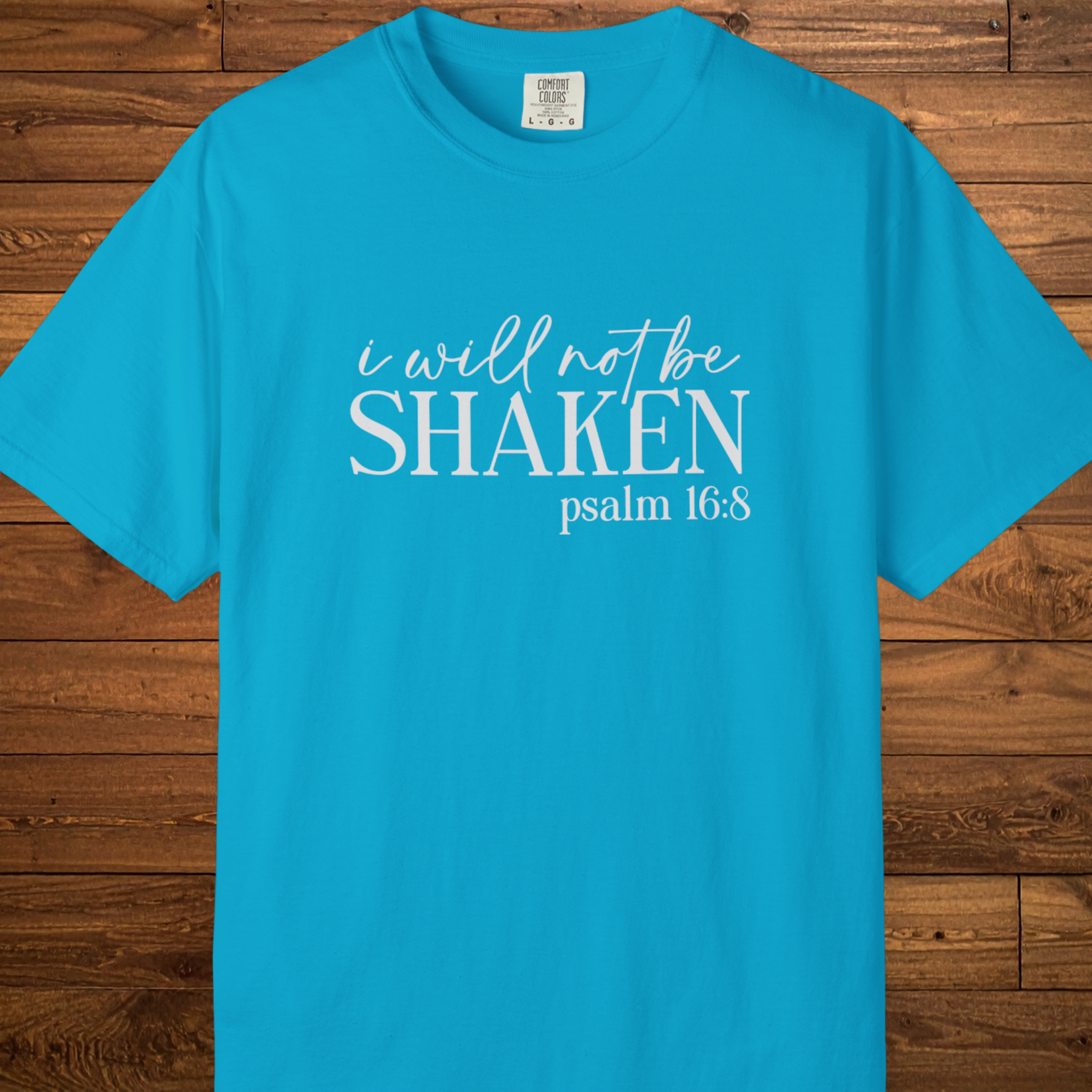 A bright turquoise blue short-sleeve t-shirt with white printed text reading 'i will not be SHAKEN psalm 16:8' laid flat against a brown wood plank background.