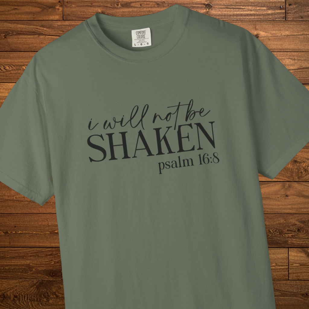 An olive green short-sleeve t-shirt with the text 'i will not be SHAKEN psalm 16:8' printed in black across the chest, displayed flat against a rustic wooden plank background.