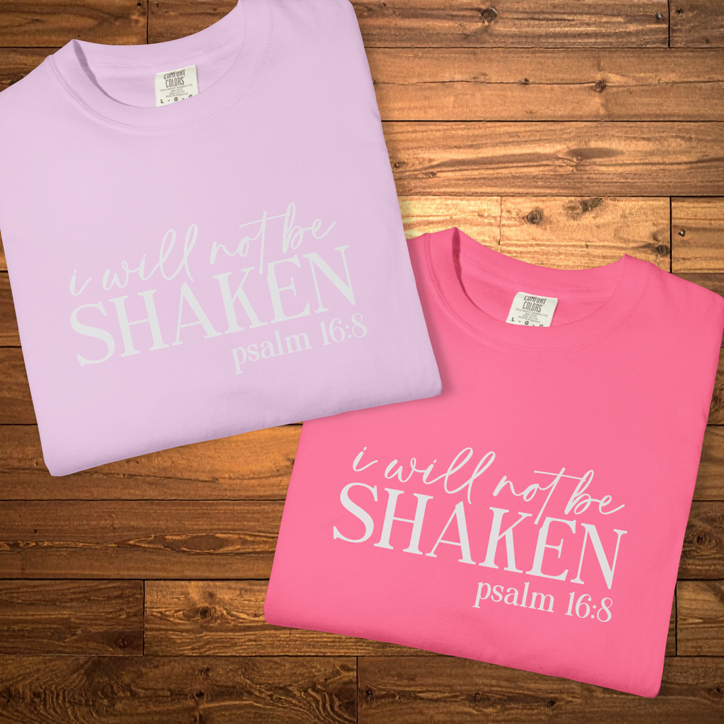 Two folded short-sleeve t-shirts with the white printed text 'i will not be SHAKEN psalm 16:8' displayed on a rustic wooden plank background. One shirt is light lavender and the other is bright pink.