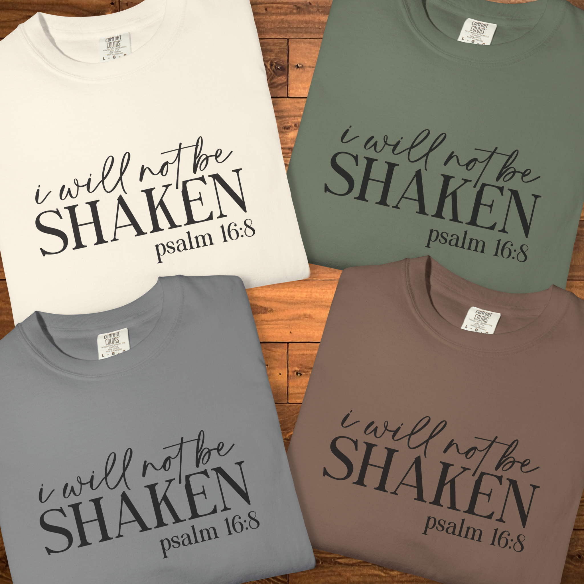Four folded short-sleeve T-shirts arranged on a wooden surface, each printed with the phrase 'i will not be SHAKEN psalm 16:8' in black script and block lettering. The shirts come in off-white, sage green, medium gray, and taupe brown.