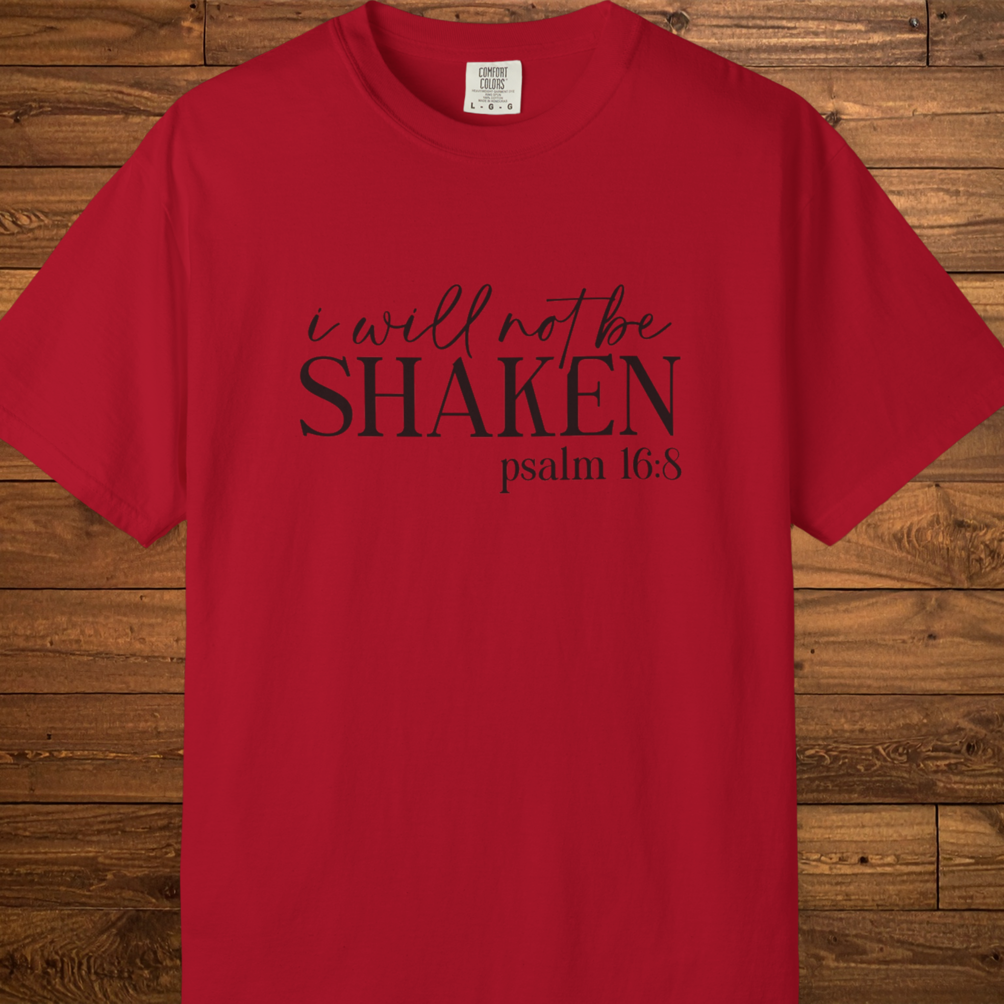 A red short-sleeve crewneck t-shirt with the text 'i will not be SHAKEN psalm 16:8' printed in black across the chest, displayed flat against a rustic wooden plank background.
