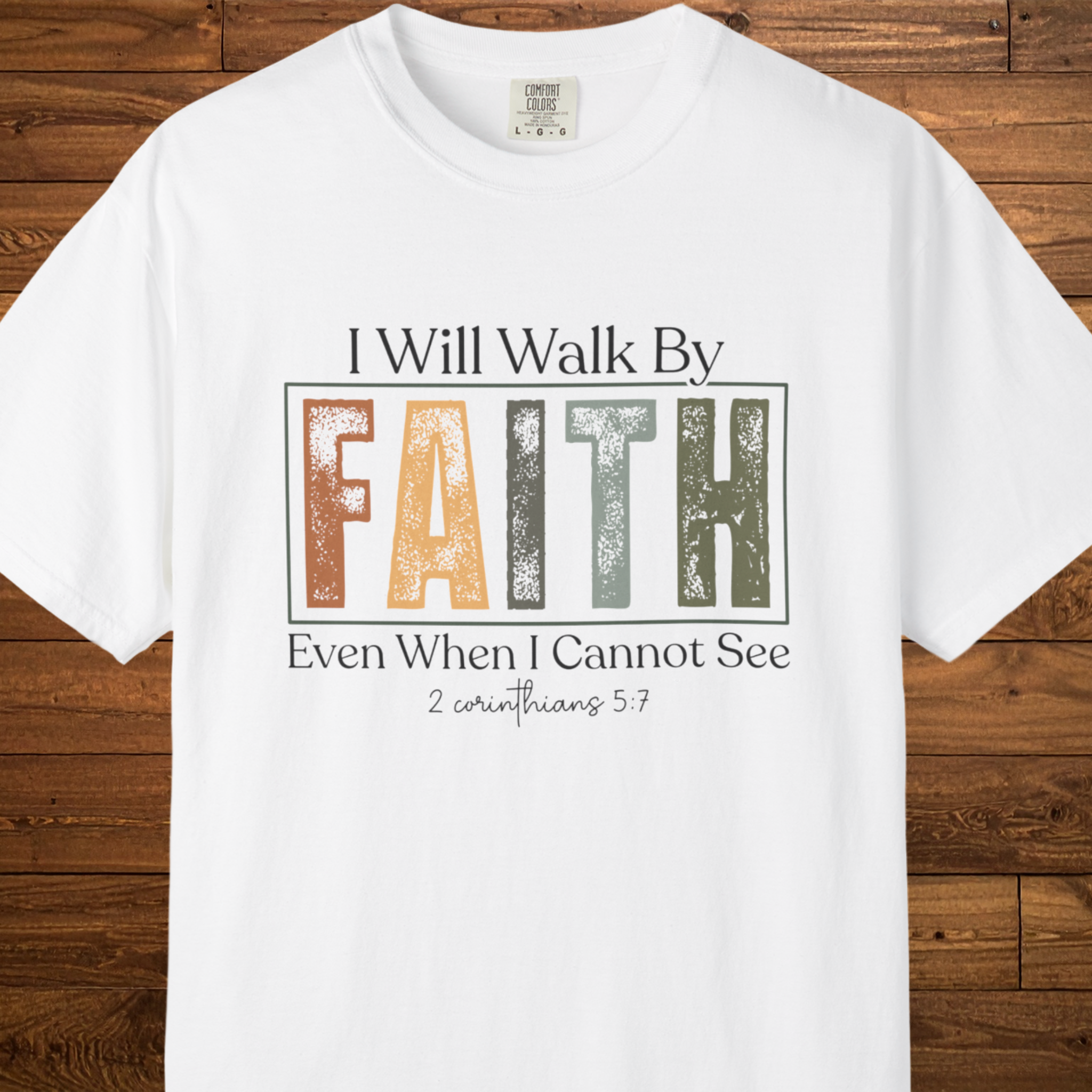 A white short-sleeve crewneck t-shirt with a graphic print on the front. The graphic reads 'I Will Walk By FAITH Even When I Cannot See' with 'FAITH' in a distressed, multi-colored block font (orange, yellow, green, gray) on a wooden plank background.