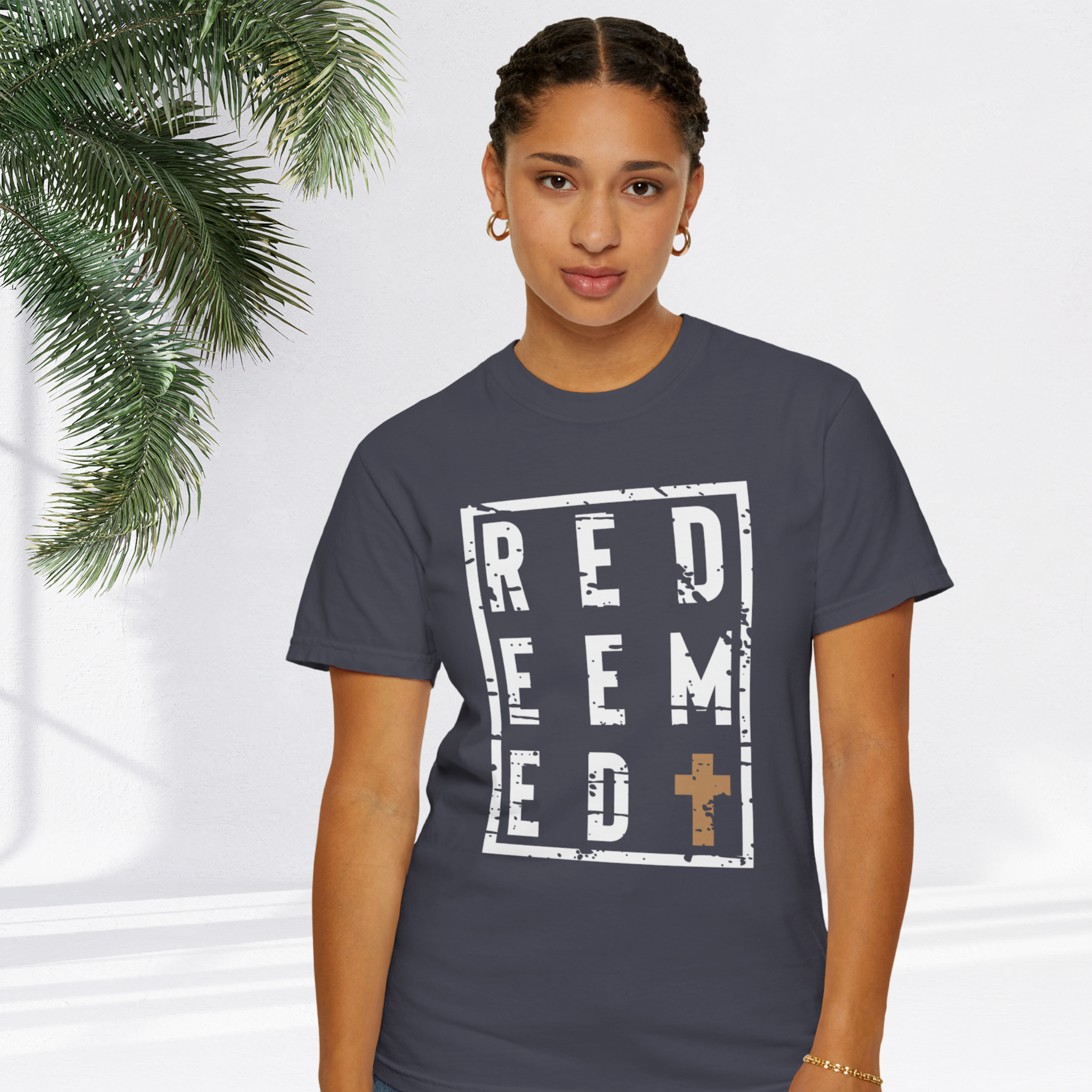 A woman wearing a dark gray t-shirt with a distressed white graphic that reads 'REDEEMED' and a small gold cross, posed against a bright white background with a palm leaf on the left.