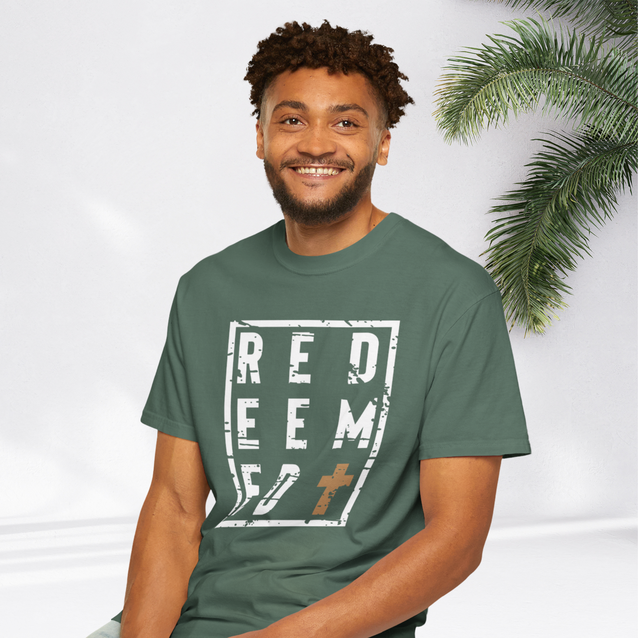 A smiling man wearing a dark green t-shirt with a distressed white graphic that reads 'REDEEMED' and a small brown cross, posed against a white background with a palm leaf on the right.
