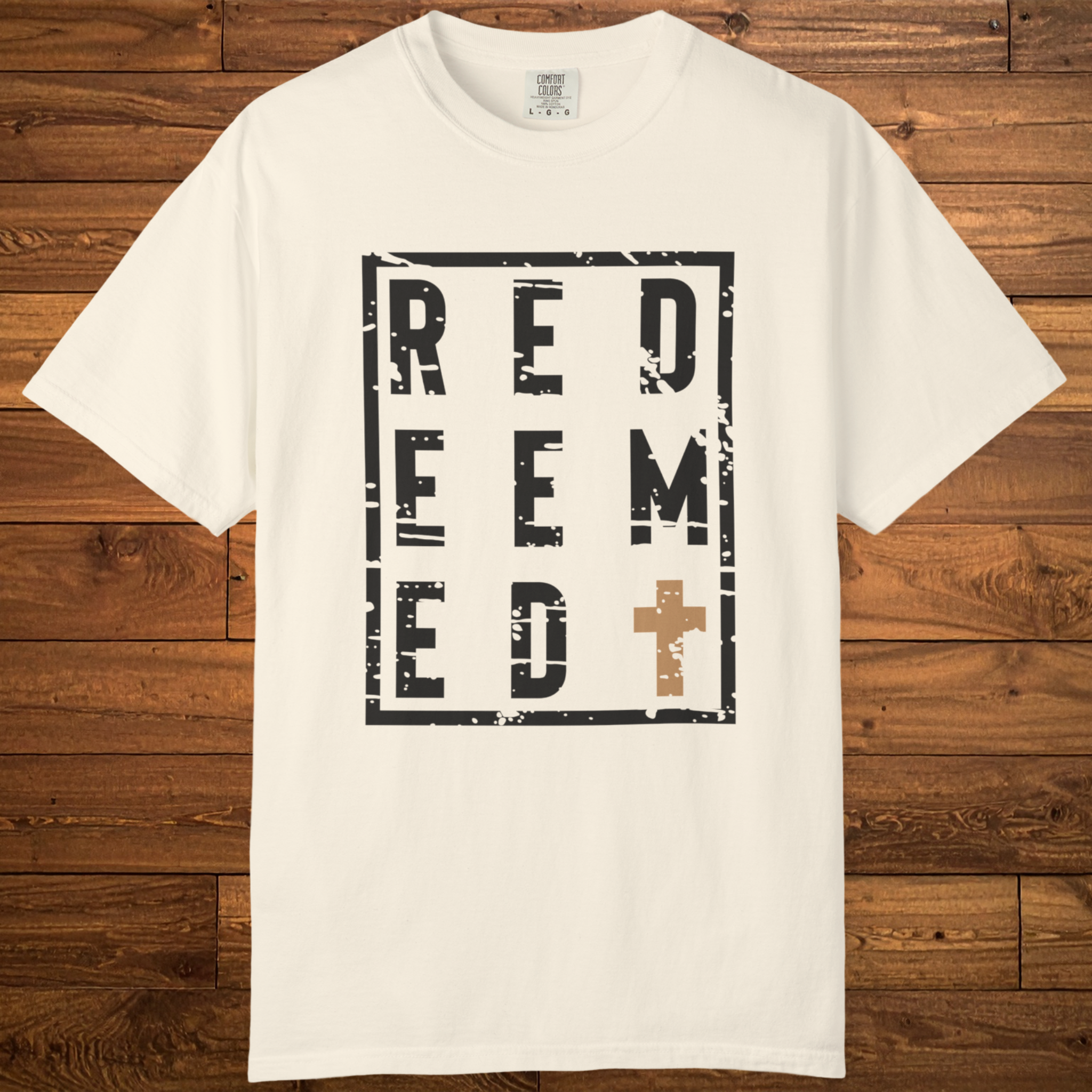 An off-white crewneck t-shirt with a distressed black graphic reading 'REDEEM' and a brown cross, laid flat on a rustic wooden background.