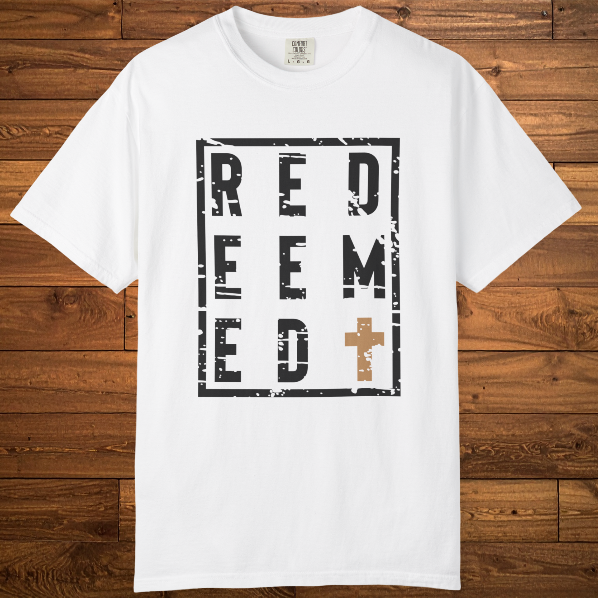 A white short-sleeve crewneck t-shirt with a distressed black graphic reading 'REDEEMED' in a square frame, featuring a brown cross on the letter 'M', displayed flat against a brown wooden plank background.