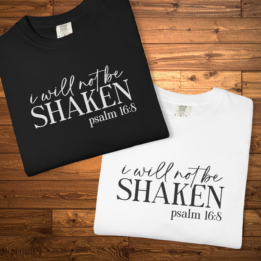 Two folded T-shirts on a wooden surface: one black with white text reading 'i will not be SHAKEN psalm 16:8' and one white with black text reading 'i will not be SHAKEN psalm 16:8'.